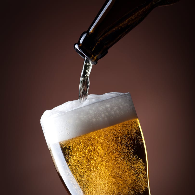 Beer pouring from a bottle into a glass, creating a frothy head against a brown background.