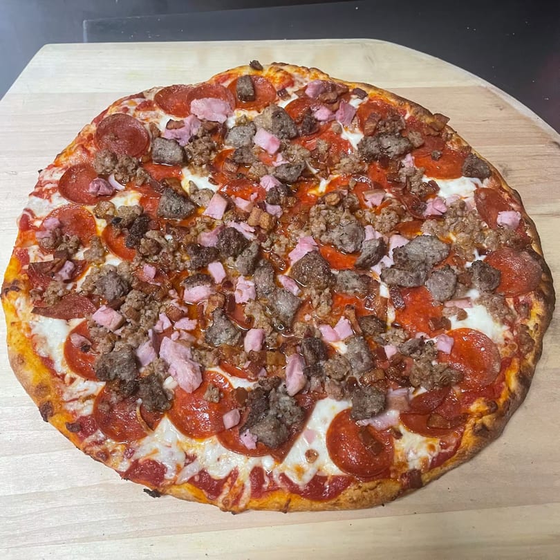 A TopCrust Jupiter Kitchen pizza, loaded with pepperoni, sausage, and ham, on a wooden peel.