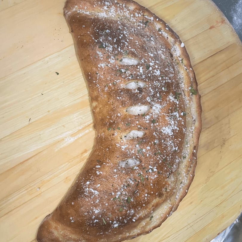 A golden-brown calzone, sprinkled with salt and herbs, rests on a wooden cutting board.